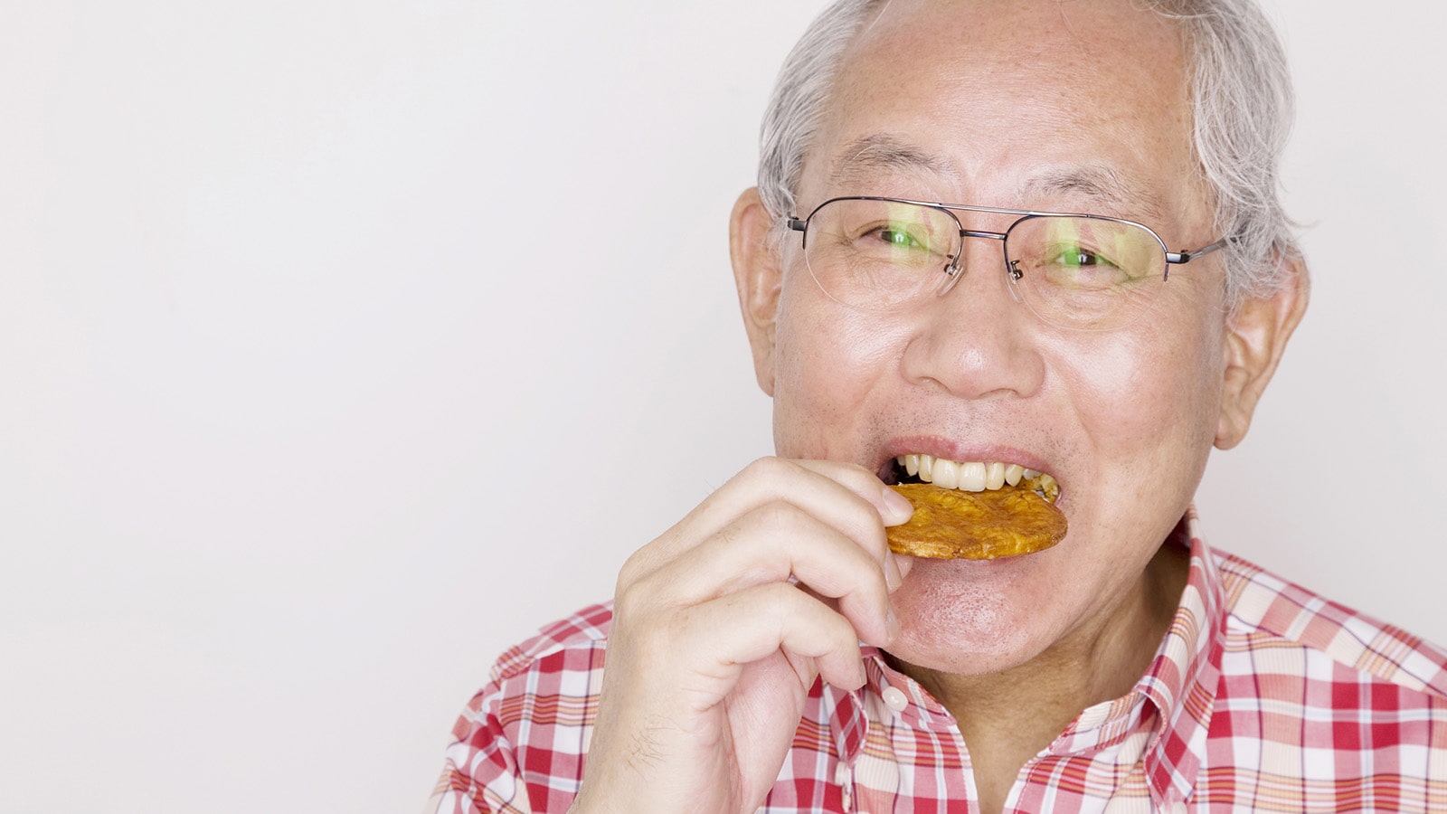 Senior asian man wearing pink plaid shirt eating a rice crackers