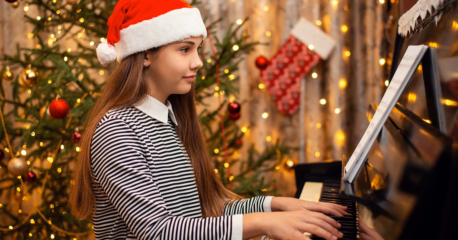 young girl wearing a santa hat playing piano in front of christmas tree