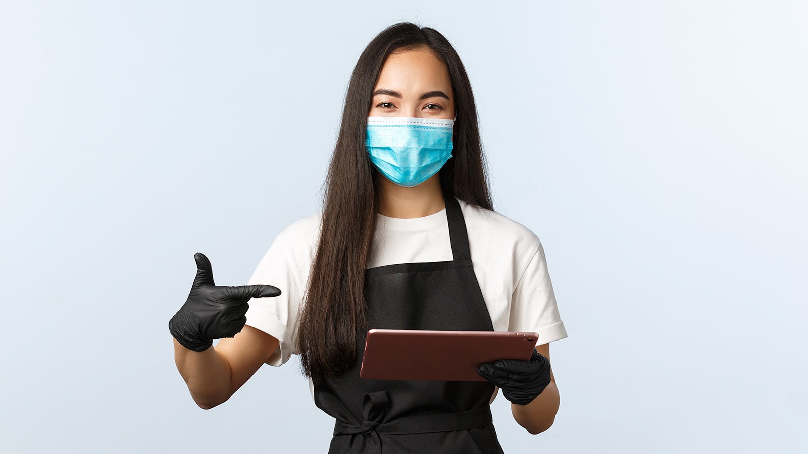 Young woman with long hair wearing face mask and apron wearing black glove and holding a tablet