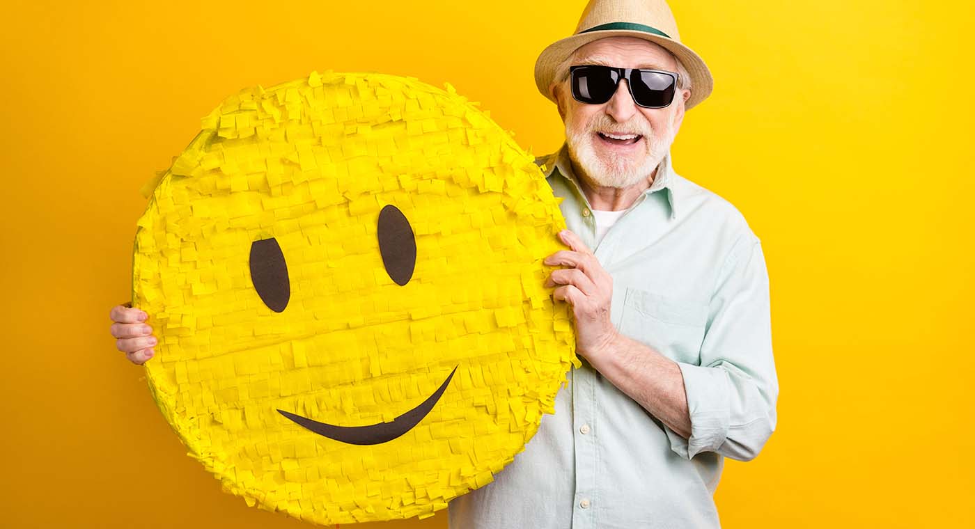 senior man wearing hat and sunglasses holding up a yellow smile face pinata in front of yellow background