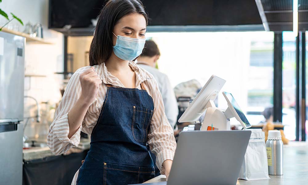 Girl in striped shirt and denim apron wearing mask using a laptop