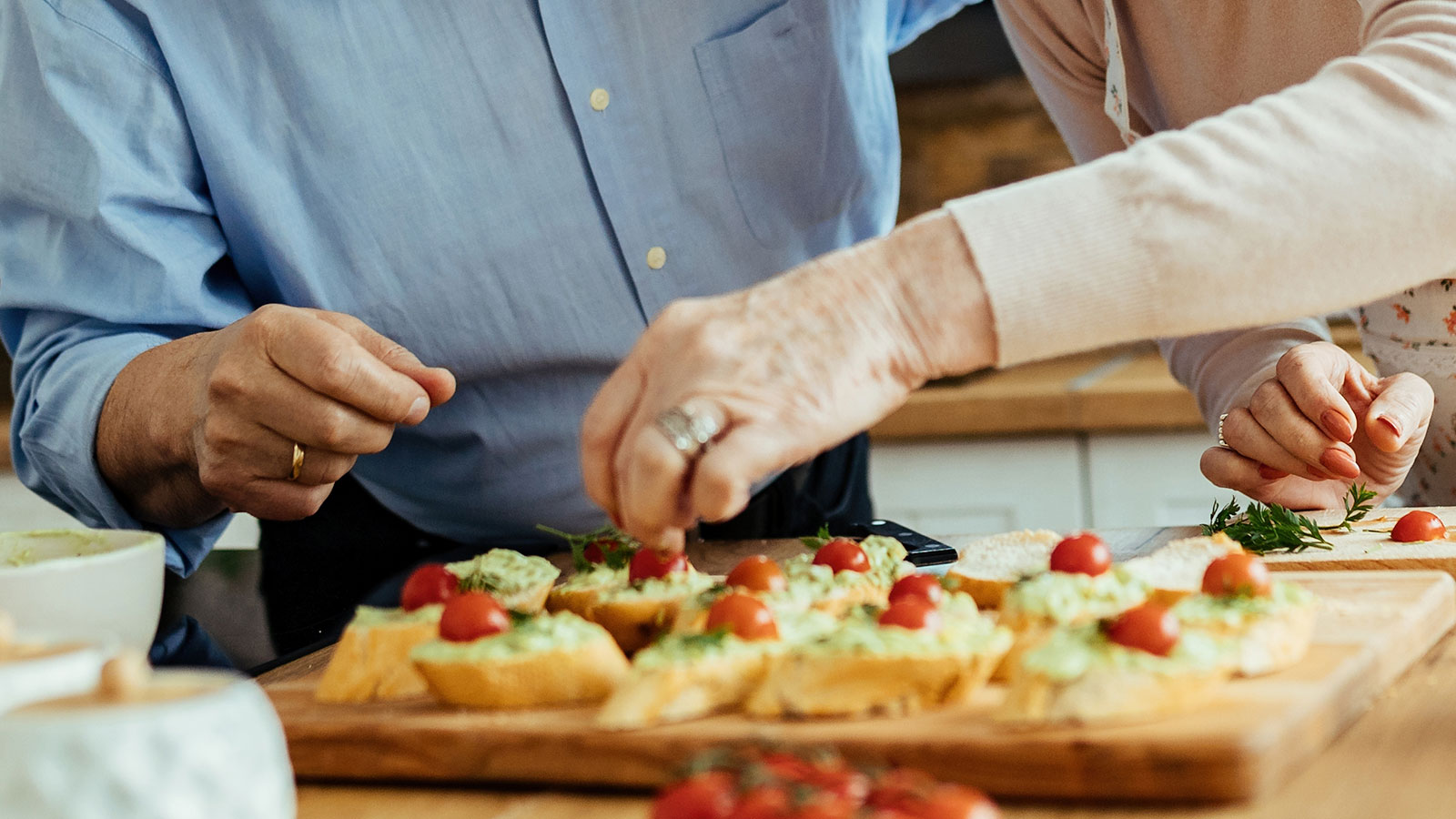 Senior man with mustache wearing a blue shirt and senior woman wearing apron preparing appetizers in kitchen