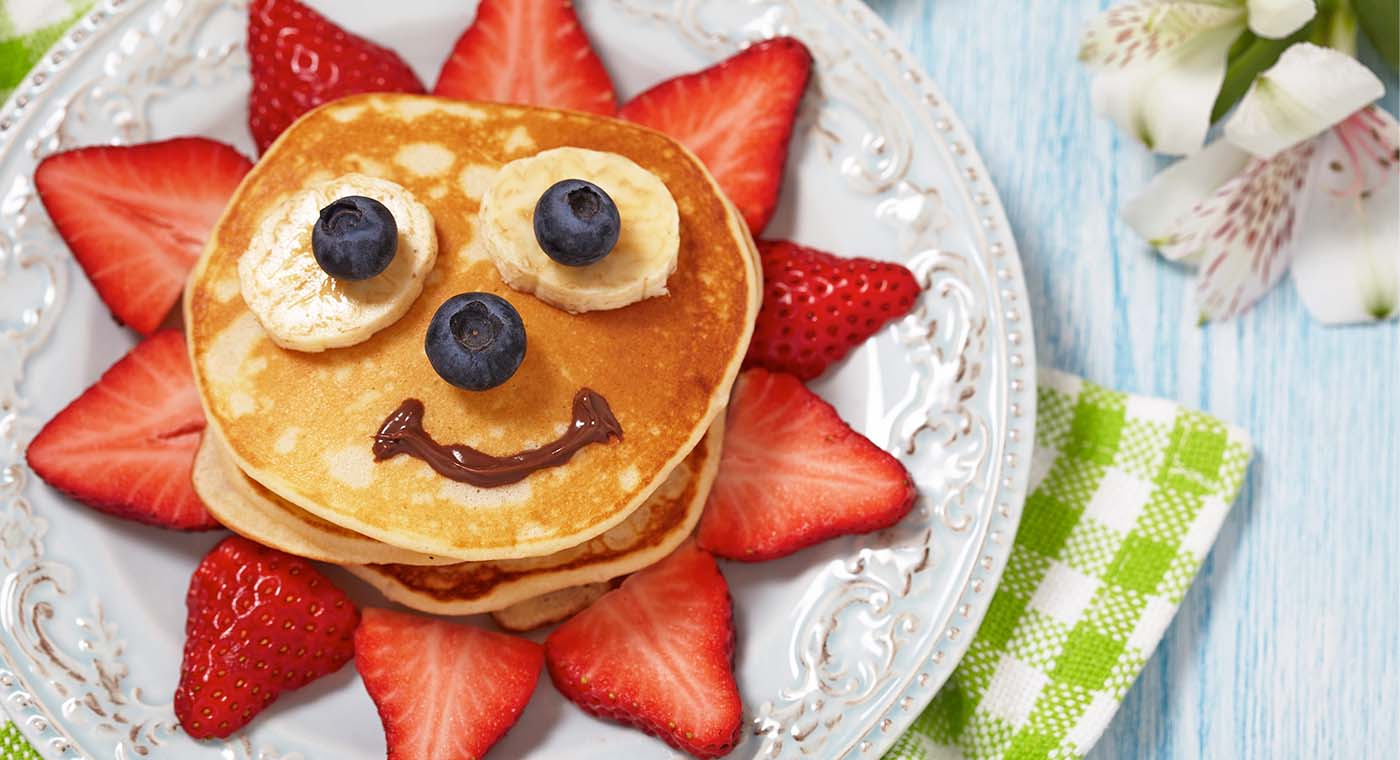 pancake breakfast with strawberries and a face made from blueberries, bananas and maple syrup