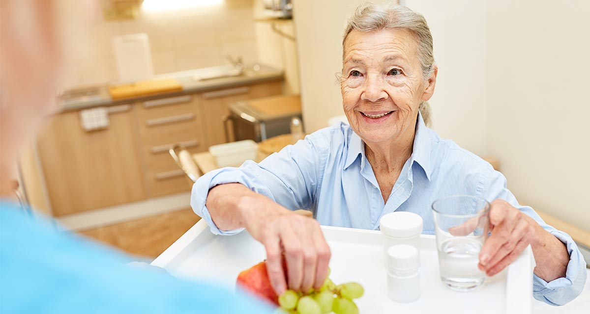 elderly woman holding a glass of water reaching for pills from a food tray