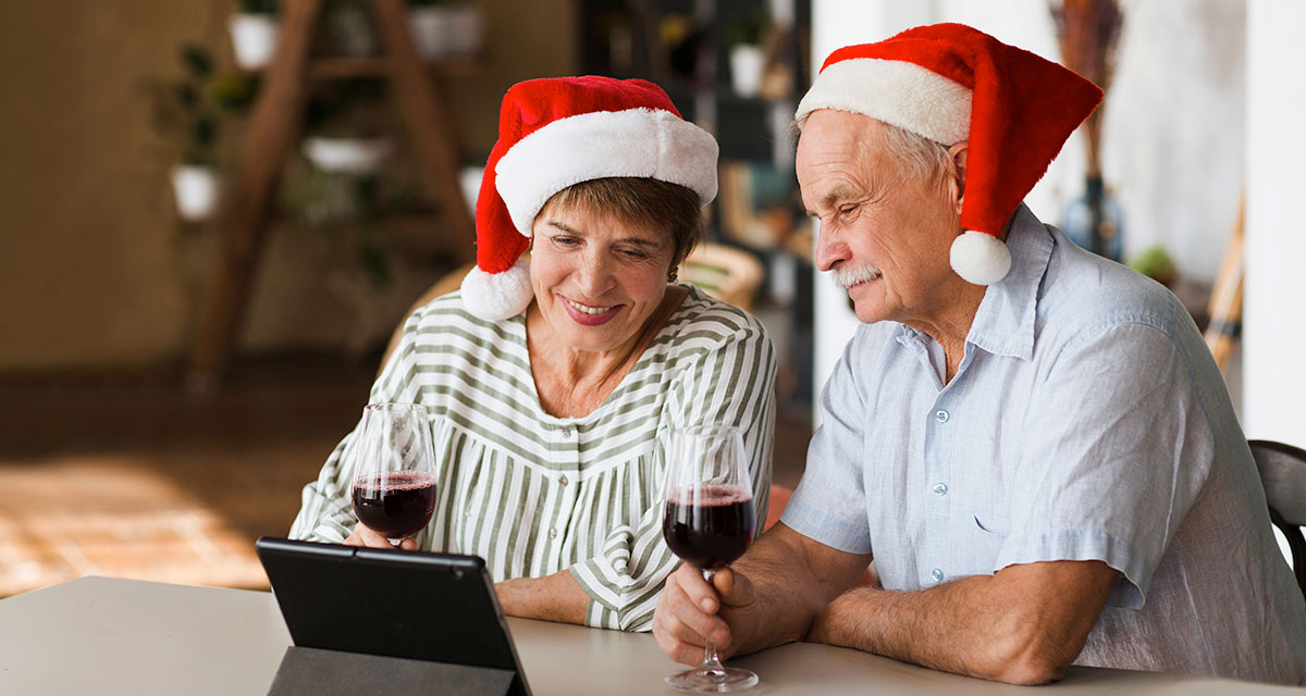 Older couple wearing santa hats at christmas looking at tablet and drinking red wine