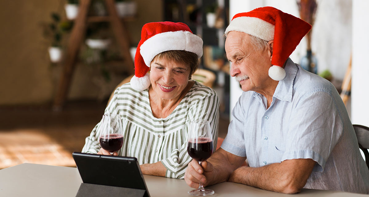 Older couple wearing santa hats at christmas looking at tablet and drinking red wine