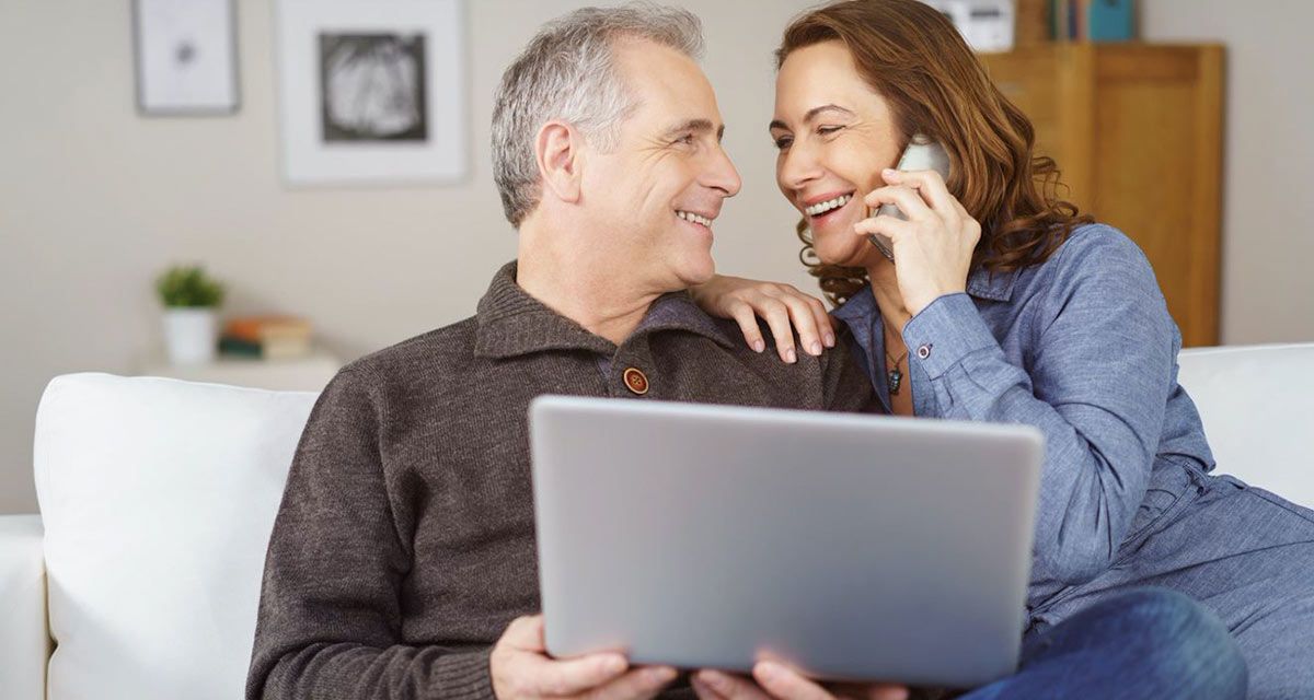 Couple smiling on couch as they look on their computer and talk to family over the phone