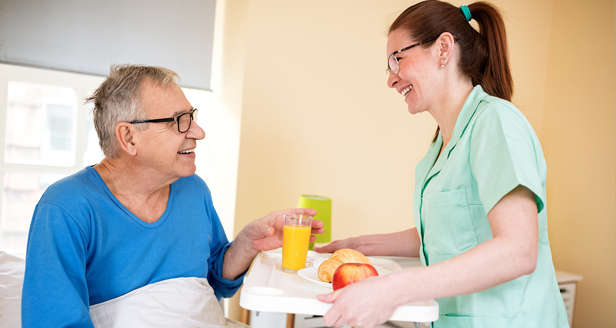 Smliling nurse wearing green serving a tray of food to happy patient wearing a blue shirt