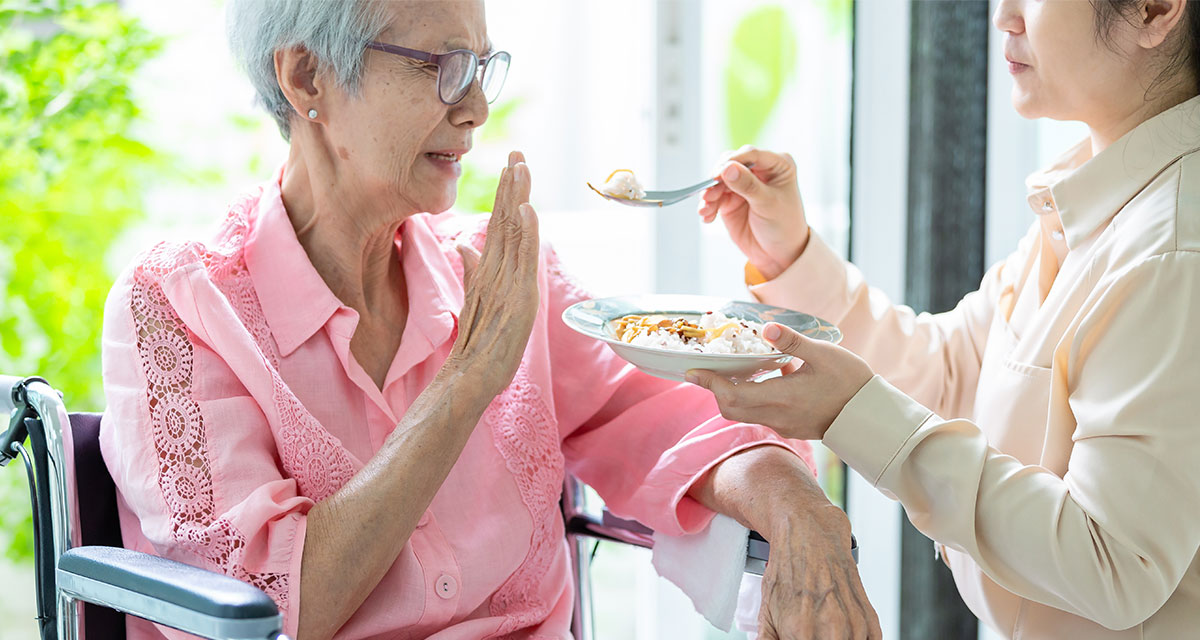 Resident at Longterm care facility in pink shirt on blue wheelchair refusing a spoonful of food from caretaker