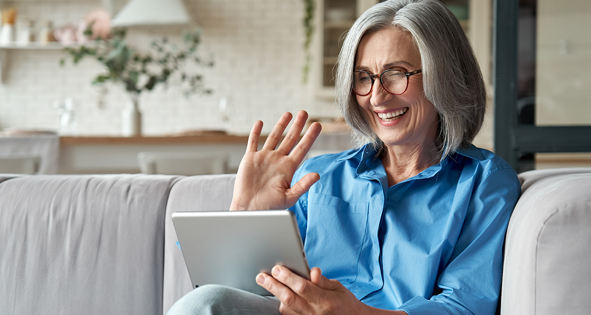 Older grey haired woman in blue shirt sitting on couch smiling while holding a tablet