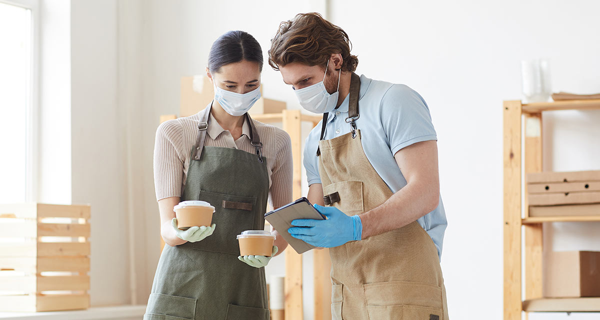 Couple in apron wearing masks holding bowls and looking at tablet