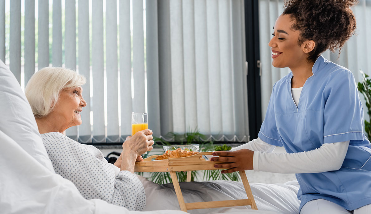 Smiling female african amercian nurse delivering food tray to smiling caucasian femail patient smiling 