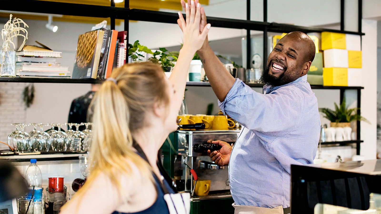 Caucasian woman with hair in ponytail giving a high five to an african american man in blue shir at a cafe