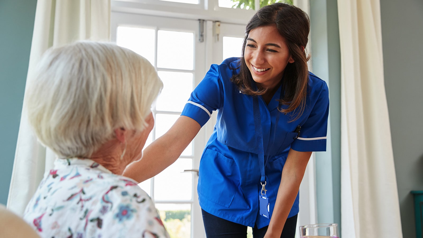 Nurse wearing blue smiling at elderly resident after delivering meal 