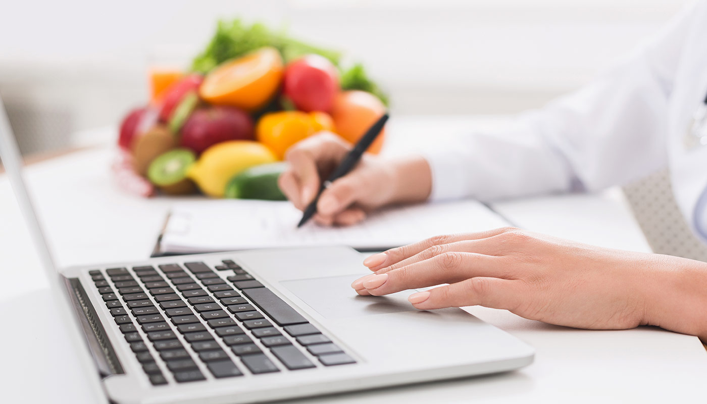 Nutritionsit using laptop and writing down notes with stack of fruit in the background