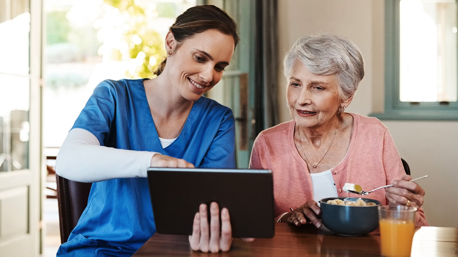 Nurse in blue smock showing elderly woman wearing pink something on a tablet while she eats breakfast
