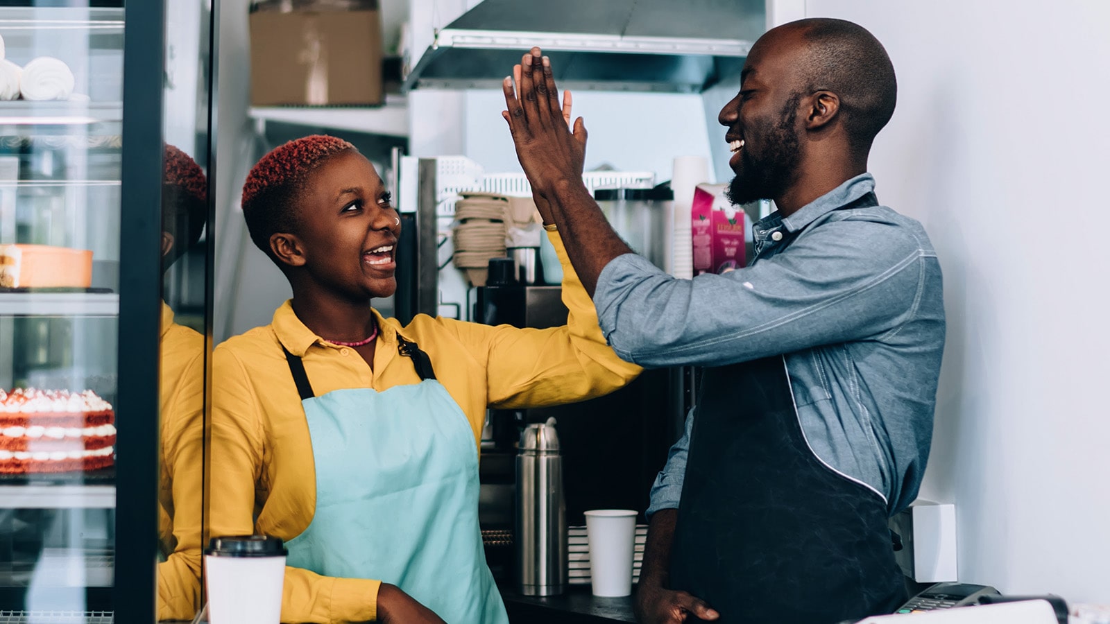 Cheerful African American man and woman in aprons giving high five to each other while celebrating success and standing behind counter in small restaurant